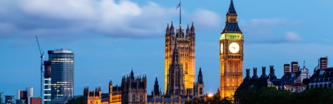 UK Parliament building in the City of London. Dusk setting with clouds in the background. Lights are lit up on Big Ben clock. English buildings in the foreground and Union Jack flag visible.