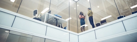 African ethinicity woman and white man standing inside a modern office building in a financial city district. Woman has long dreads and is wearing a skirt with a red floral top. Colleagues talking to each other near desks with computers and chairs. Manager and employee stood near glass windows on high story building communicating and smiling.