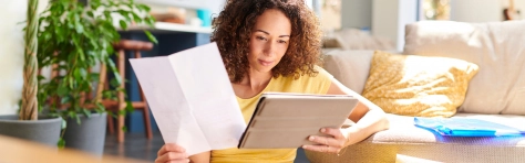 Image of a woman holding a piece of paper in one hand and a tablet in the other, checking her insurance online. 
