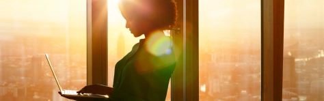 An image of a businesswoman on a laptop standing in front of a window with the sun shining in 
