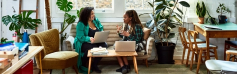 Two African black women sitting down on sofa at home in living room. Adults discussing finances with laptops and paperwork on the table. Quirky and modern interior home with wooden chairs and house plants.