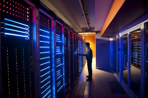 Male software engineer standing next to super computers / data software centre assessing equipment. Dark room with high power technology for commercial use. 